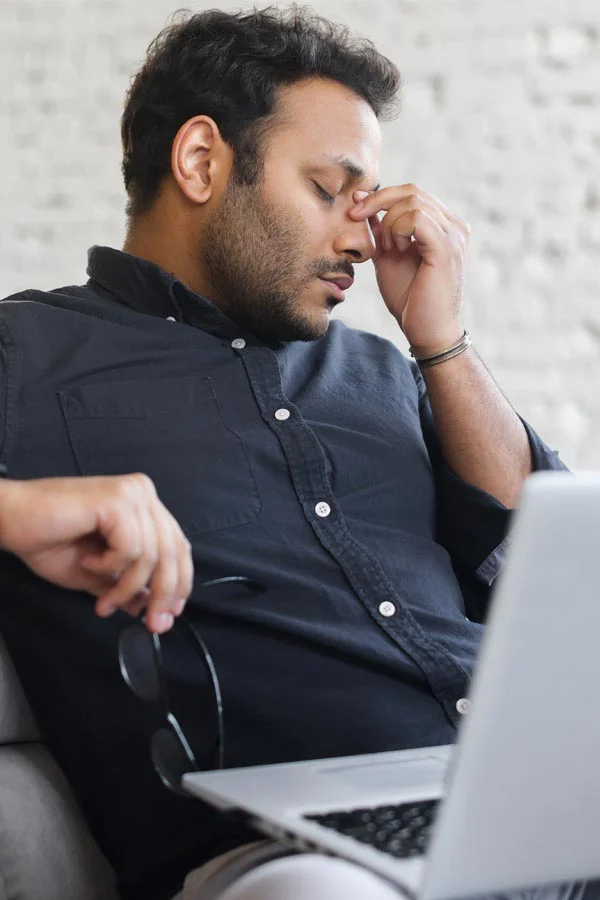A middle-aged man with a laptop closing his eyes and rubbing his nose. He needs treatment for Adrenal Fatigue Syndrome from Yashoda Singh, MD in Hamilton-Toronto-Niagara.