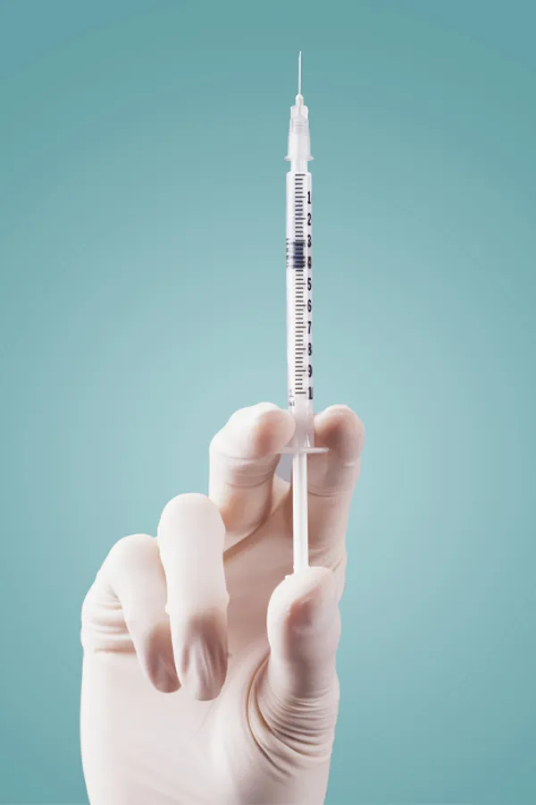 Close-up of a white-gloved hand holding a syringe against a light blue background, representing aesthetic filler services from Yashoda Singh, MD in Hamilton-Toronto-Niagara.