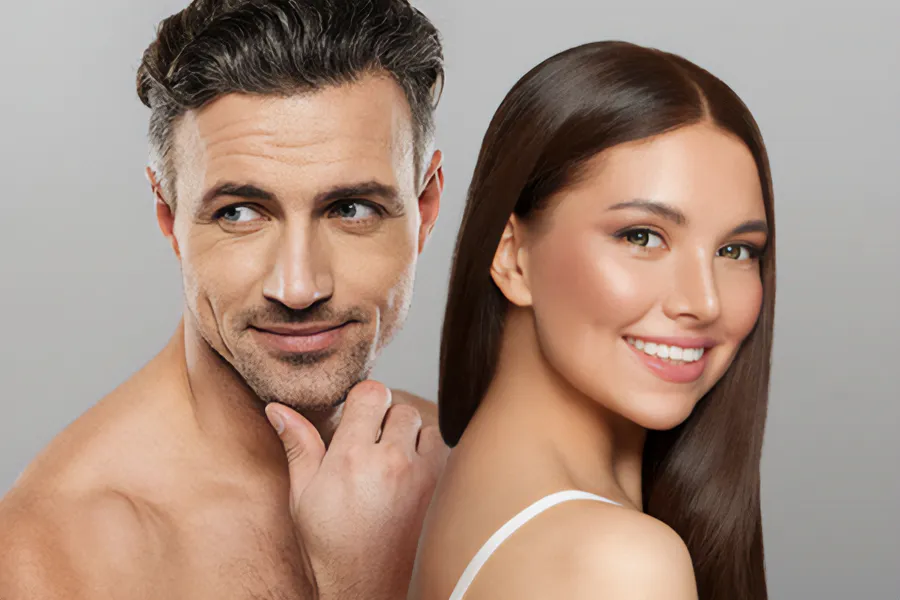 A handsome man and beautiful brunette woman stand against a gray background showing their smooth beautiful skin resulting from aesthetic and cosmetic care from Yashoda Singh, MD in Hamilton-Toronto-Niagara.