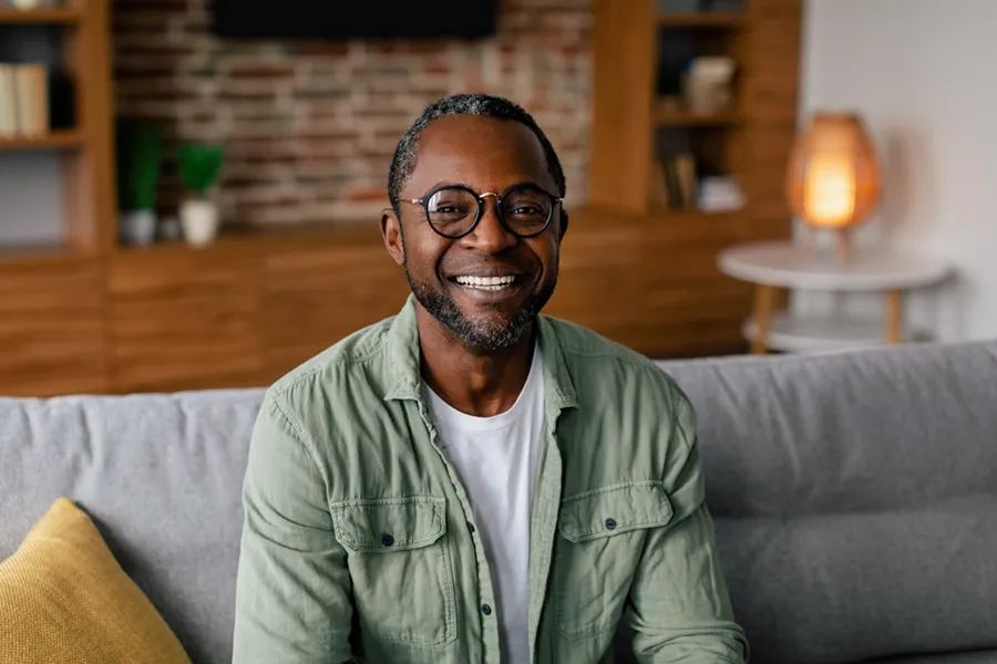 A middle aged black man in a green button up shirt sits on his couch smiling, happy with his treatment for andropause and low testosterone from Yashoda Singh, MD in Hamilton-Toronto-Niagara.