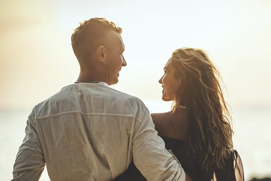 A couple gazes at each other on the beach at sunrise, viewed from behind. Schedule Anti-Aging treatment from Yashoda Singh, MD in Hamilton-Toronto-Niagara.
