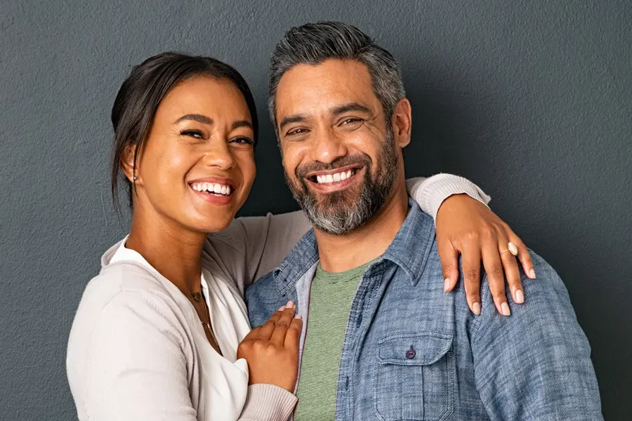 A middle-aged couple with their arms around each other standing against a gray background and smiling, happy with the bioidentical hormone therapy they received from Yashoda Singh, MD in Hamilton-Toronto-Niagara.