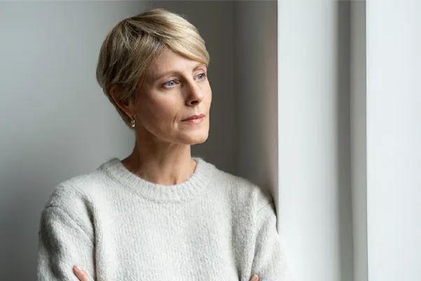 A blond, middle-aged woman looks out the window with a tired expression. Get chronic fatigue treatment from Yashoda Singh, MD in Hamilton-Toronto-Niagara.