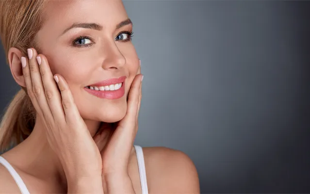A middle-aged blonde woman with beautiful skin smiles and holds her face. Get facial treatments from Yashoda Singh, MD in Hamilton-Toronto-Niagara.