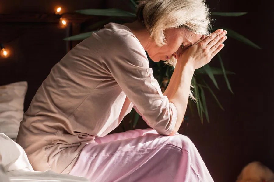 A woman sitting on the edge of her bed holds her head in her hands. Schedule hormonal mental health treatment from Yashoda Singh, MD in Hamilton-Toronto-Niagara.