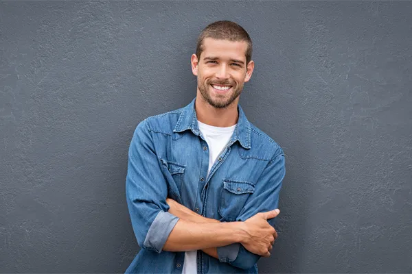 A man in a denim shirt stands smiling against a gray-blue wall, pleased with his testosterone hormone therapy from Yashoda Singh, MD in Hamilton-Toronto-Niagara.