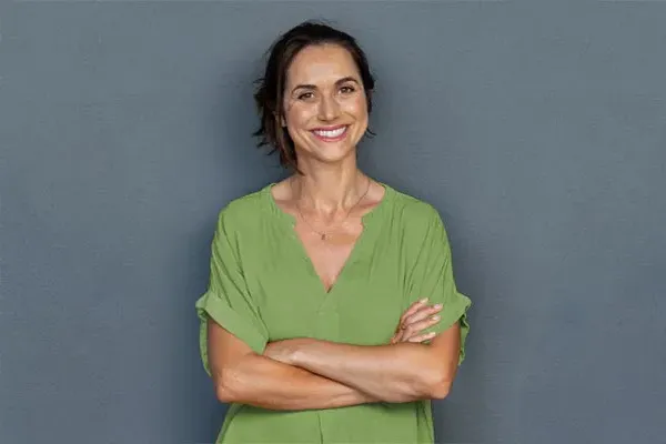 A woman in a green shirt stands smiling against a gray-blue wall, pleased with her hormone therapy from Yashoda Singh, MD in Hamilton-Toronto-Niagara.