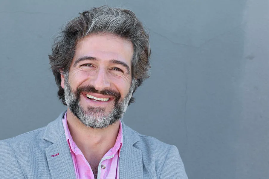 A smiling man with salt and pepper hair and beard wearing a pink shirt and gray blazer stands agains a gray wall. Get hypogonadism treatment from Yashoda Singh, MD in Hamilton-Toronto-Niagara.