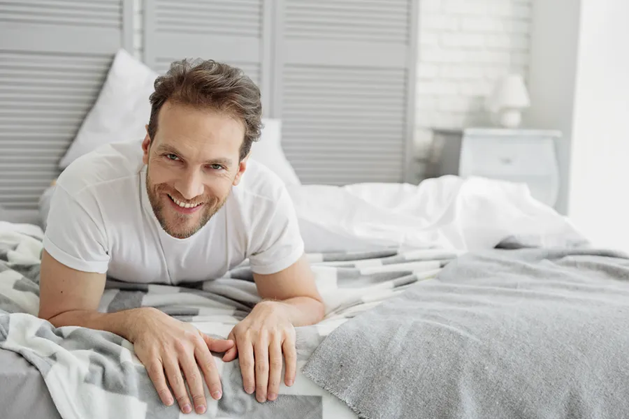 A dirty-blonde haired man lays on his stomach in bed smiling. Schedule treatment for low libido in men from Yashoda Singh, MD in Hamilton-Toronto-Niagara.