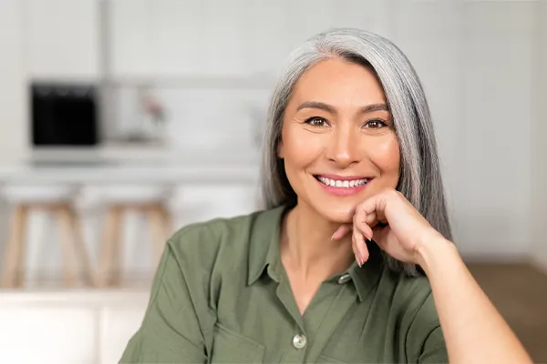 An asian woman with long gray hair and a green shirt sitting and smiling on her couch, happy with her menopause care from [NAME} in Hamilton-Toronto-Niagara.