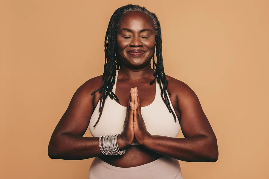 An older African American woman confidently posing against a warm background smiling with hands in a prayer pose. Yashoda Singh, MD treats obesity using functional medicine in Hamilton-Toronto-Niagara.