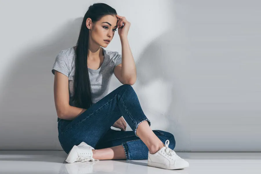 A young brunette woman in a gray t-shirt and jeans sits against a gray wall with her head resting on her hand and a distressed expression. Get help for Polycystic Ovarian Syndrome (PCOS) from Yashoda Singh, MD in Hamilton-Toronto-Niagara.