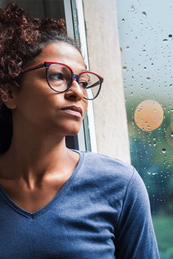 A black woman with with a somber expression and glasses and a gray shirt stares out the window on a rainy day. Get treatment for Premenstrual Dysphoric Disorder from Yashoda Singh, MD in Hamilton-Toronto-Niagara.