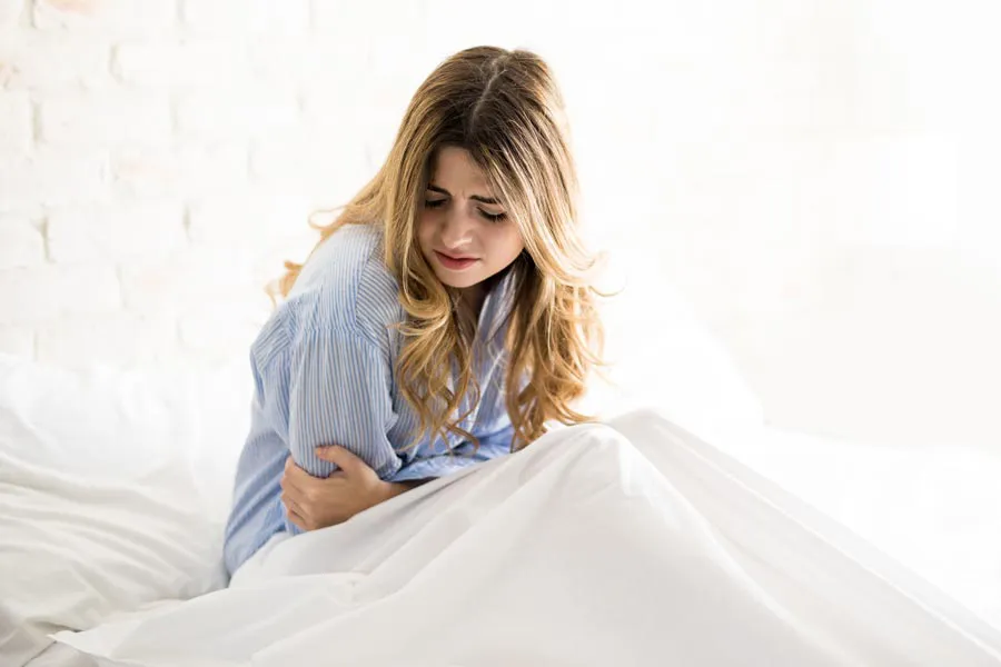 A woman with long blonde hair sits in her bed holding her stomach in pain, considering getting care for PMS from Yashoda Singh, MD in Hamilton-Toronto-Niagara.