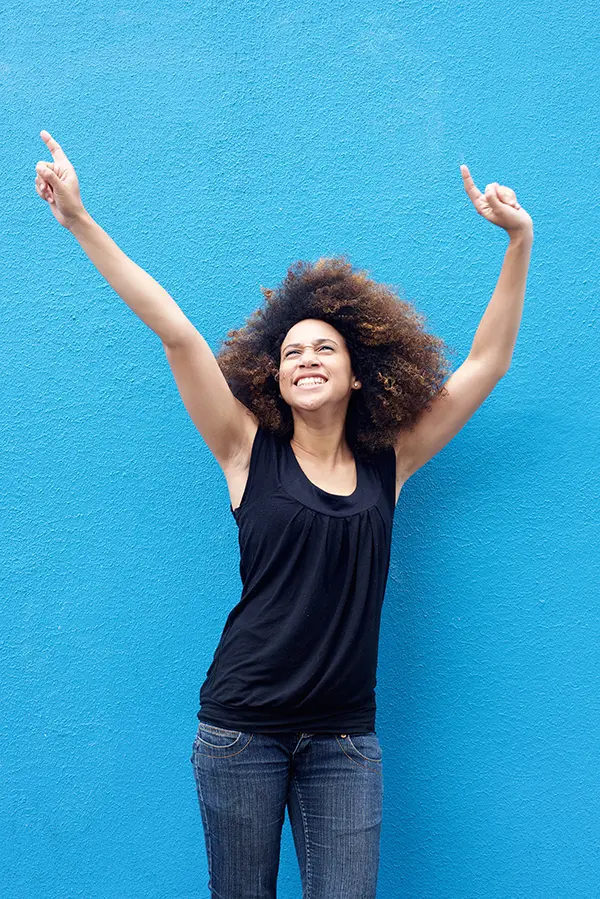 A woman in a dark blue tank top standing in front of a bright blue wall, raising her arms in celebration of relief from PMS from Yashoda Singh, MD in Hamilton-Toronto-Niagara.