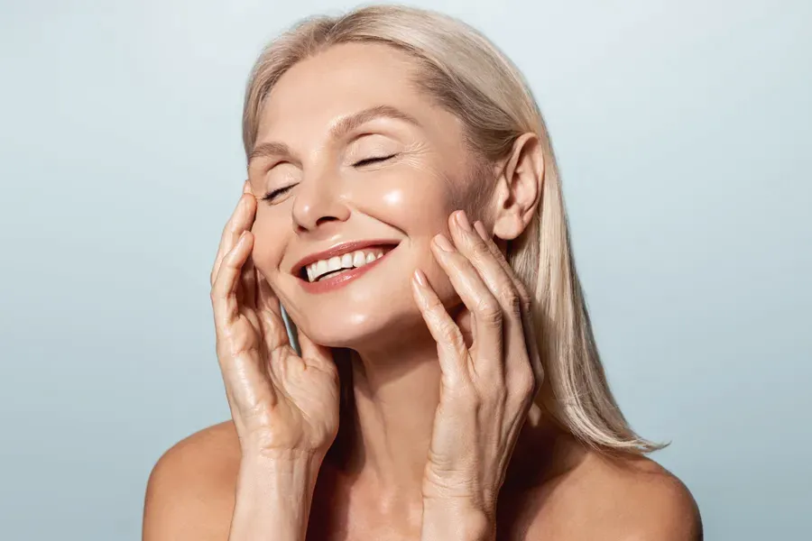 A beautiful mature blonde woman touches her face, happy with her PRP facelift and facial injections from Yashoda Singh, MD in Hamilton-Toronto-Niagara.