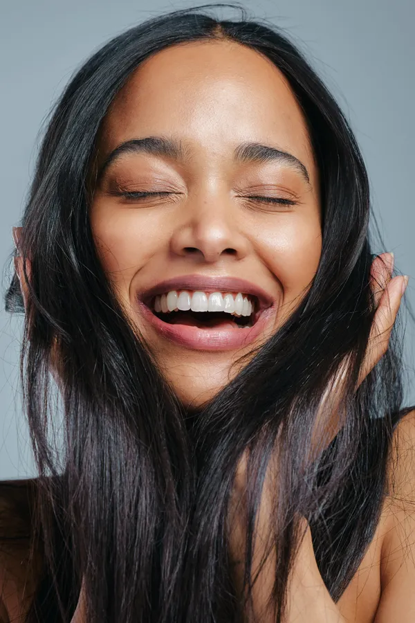 A beautiful latin woman smiles and runs her fingers through her long, beautiful hair, happy with her PRP for hair loss and restoration treatment from Yashoda Singh, MD in Hamilton-Toronto-Niagara.