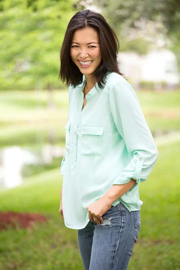 A middle-aged brunette woman in a light green button-up shirt stands outside smiling, happy with her perimenopause treatment from Yashoda Singh, MD in Hamilton-Toronto-Niagara.