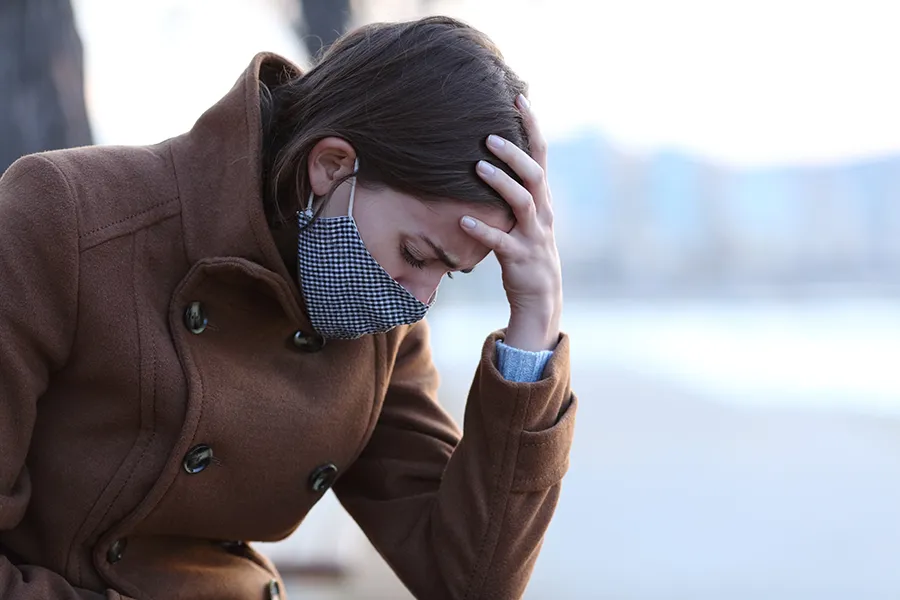 A woman in a mask sits outside with her hand on her forehead in pain. Get long covid treatment from Yashoda Singh, MD in Hamilton-Toronto-Niagara.