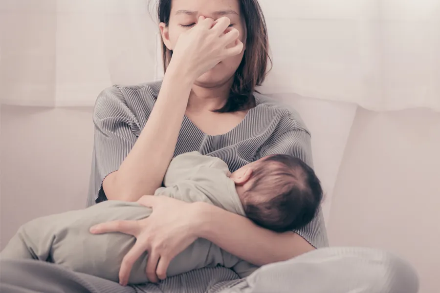 A woman nursing an infant on couch rubs her eyes with exhaustion. Get treatment for postpartum hormone imbalance from Yashoda Singh, MD in Hamilton-Toronto-Niagara.