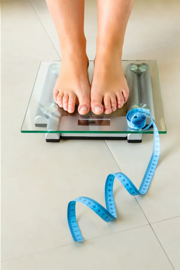 Close-up of a woman's feet standing on a scale, with measuring tape by her toes, getting treatment for weight loss resistance from Yashoda Singh, MD in Hamilton-Toronto-Niagara.